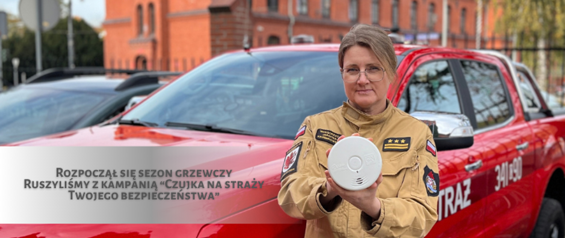 Kobieta w piaskowym mundurze strażackim prezentuje białą, okrągłą czujkę dymu na tle czerwonego wozu strażackiego. Po lewej stronie znajduje się napis: „Rozpoczął się sezon grzewczy. Ruszyliśmy z kampanią Czujka na straży Twojego bezpieczeństwa.