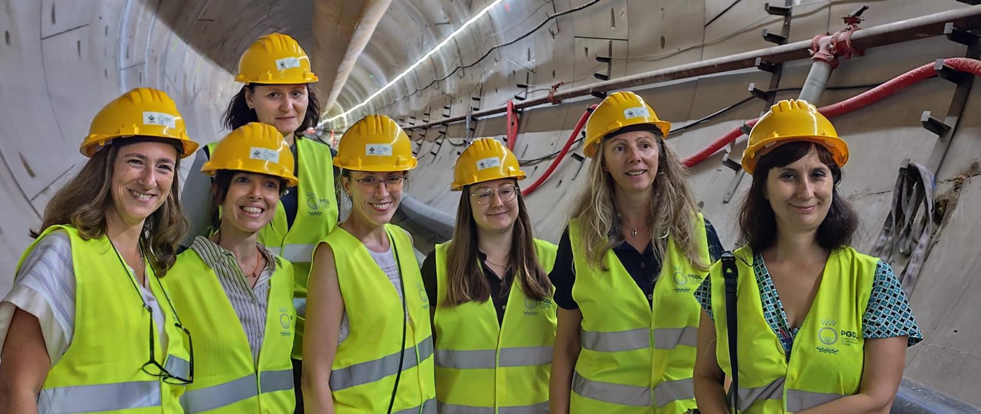 Grupa siedmiu kobiet w żółtych kaskach ochronnych i odblaskowych kamizelkach stoi wewnątrz dużego tunelu technicznego podczas wizyty na budowie. Tunel ma betonowe ściany, a pod jego stropem biegnie duża rura. Wzdłuż ścian widoczne są przewody i elementy instalacji. Zdjęcie wykonano z perspektywy podkreślającej długość i głębokość tunelu. Oświetlenie jest sztuczne, a dominujące kolory to szarość betonu i jaskrawa żółć kamizelek oraz kasków.