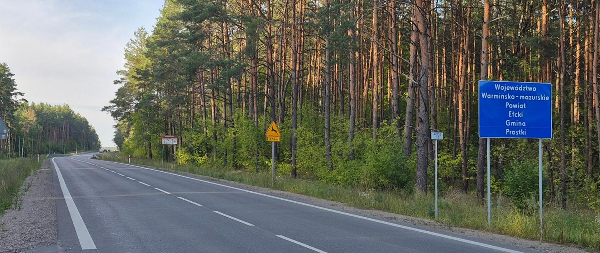 Droga krajowa nr 65 biegnąca przez teren leśny. Po prawej stronie jezdni widoczna jest niebieska tablica informacyjna z napisem „Województwo Warmińsko-Mazurskie, Powiat Ełcki, Gmina Prostki”, oznaczająca granicę administracyjną. Wzdłuż drogi rośnie gęsty las sosnowy.