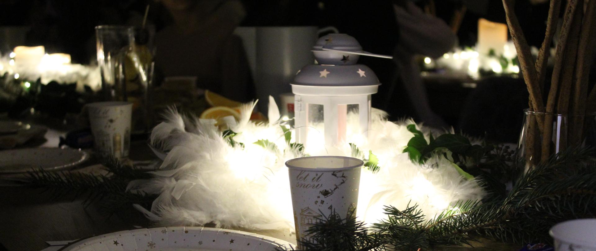 Zdjęcie ze spotkania wigilijnego przedstawia stół świąteczny udekorowany i nakryty. W środkowej części stoi zapalony lampion otoczony gałązką jodły i dekoracją z białych piór. Obok kubeczek i talerzyk. Zdjęcie jest nastrojowe, świąteczne.
