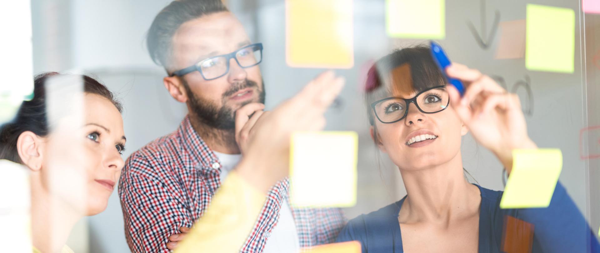 pracownicy (dwie kobiety i mężczyzna) przy tablicy zapisują pomysły