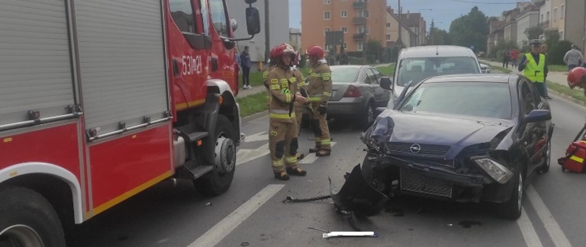 W tle stoi samochód strażacki, obok stoi jeden z rozbitych pojazdów. 