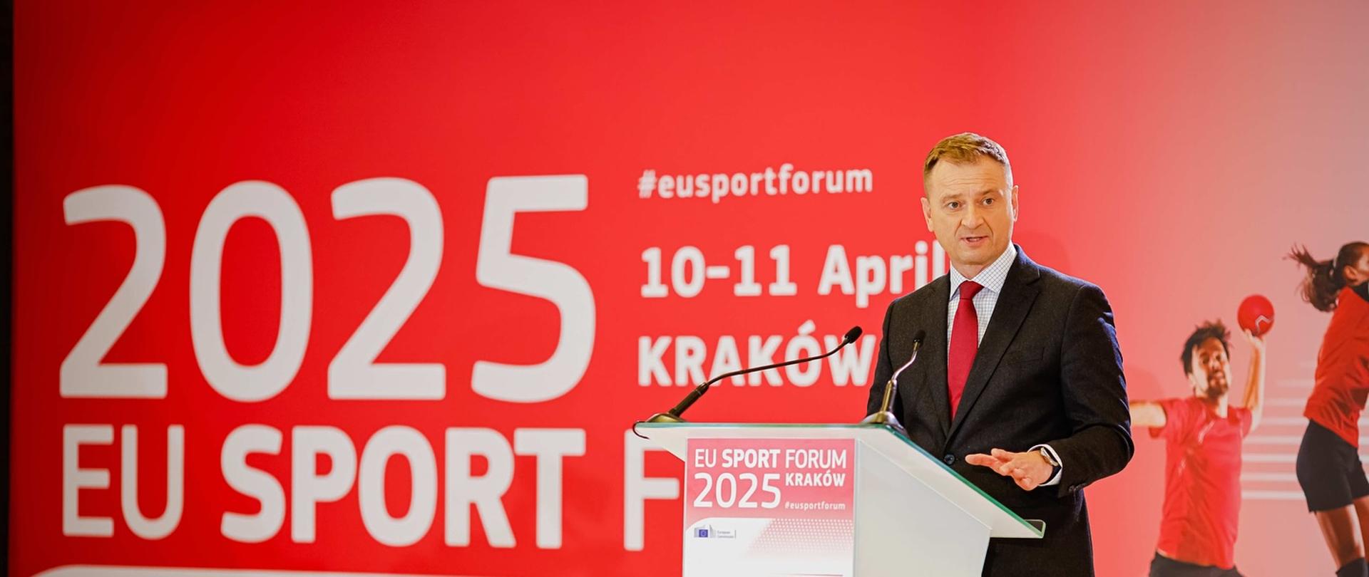 Minister Sławomir Nitras otwierający forum sportu w Krakowie. Stoi za mikrofonem na tle ekranu z logiem wydarzenia