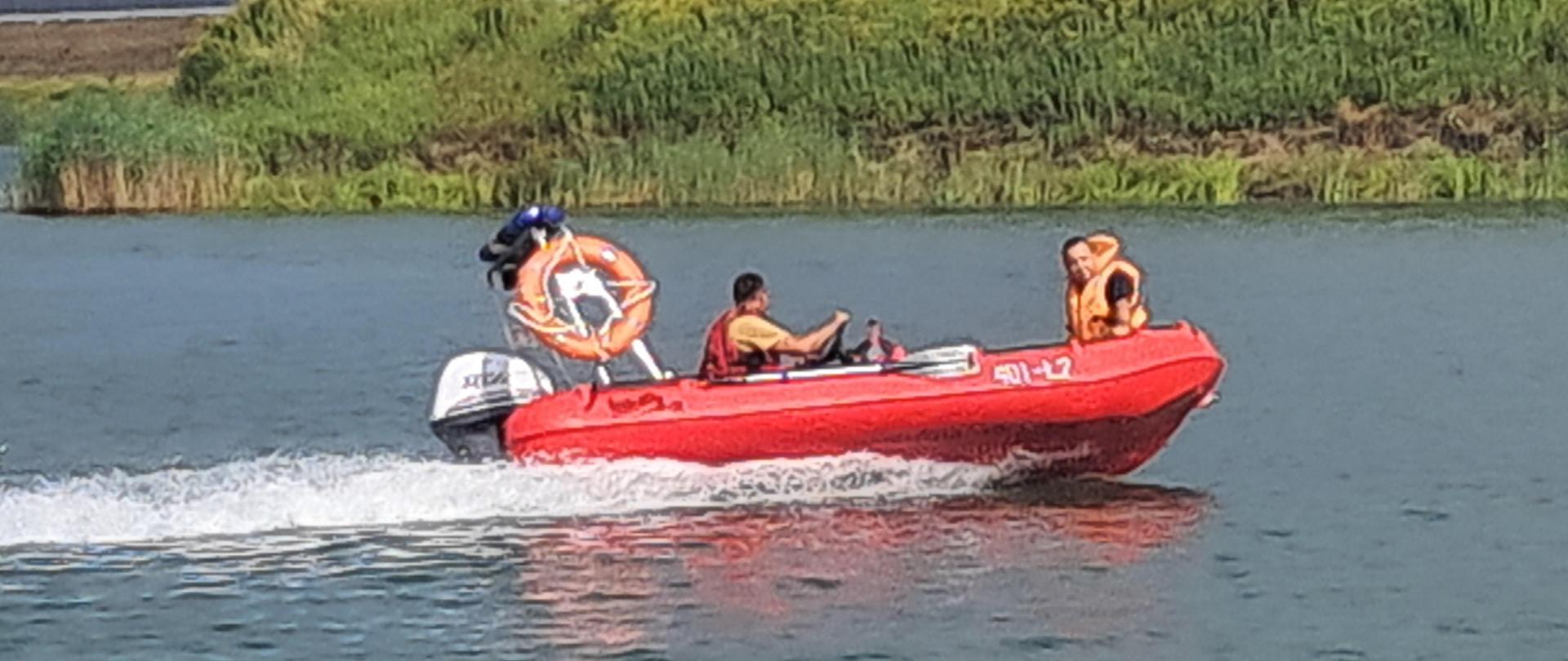 Zdjęcie przedstawia ćwiczenia z zakresu ratownictwa wodnego. Na zdjęciu widać dwóch strażaków ubranych w kamizelki ratunkowe płynących po akwenie wodnym czerwoną łodzią ratowniczą z silnikiem. Łódź ma w tylnej części przymocowaną aluminiową ramę na której znajduje się belka ze światłami błyskowymi w kolorze niebieskim. Z boku belki po prawej stronie przymocowane jest pomarańczowe koło ratownicze a po lewej stronie rzutka ratownicza. 