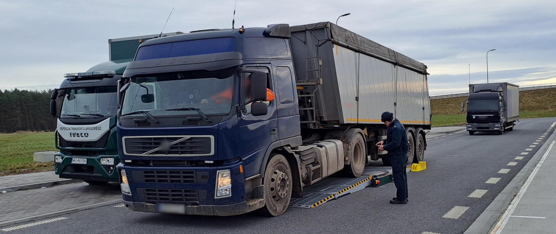 Na pierwszym planie (od prawej): umundurowany inspektor opolskiej Inspekcji Transportu Drogowego stoi obok ciągnika siodłowego z naczepą-wywrotką. Zestaw znajduje się na urządzeniach kontrolno-pomiarowych Mobilnej Jednostki Diagnostycznej. W tle: ciężarówka stoi w punkcie kontrolnym i oczekuje na szczegółową kontrolę stanu technicznego.