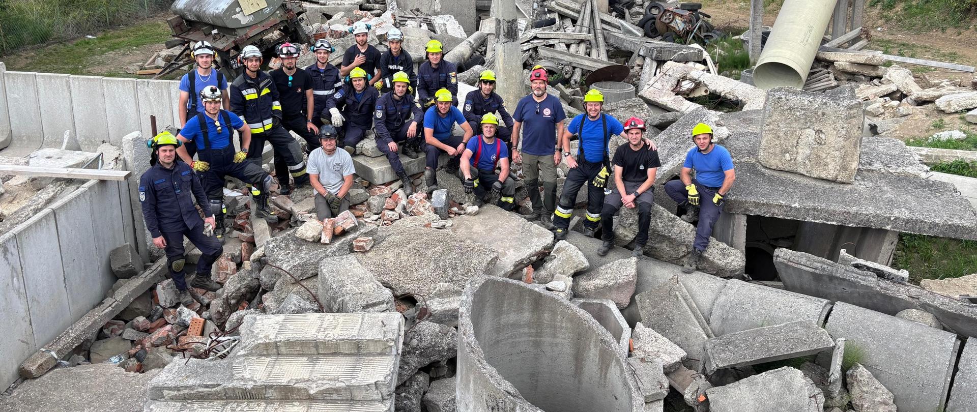 Grupa ratowników w kaskach ochronnych i umundurowaniu pozuje do wspólnego zdjęcia na tle symulowanego rumowiska powstałego po zawaleniu się konstrukcji. Otaczają ich betonowe płyty, gruzy i fragmenty budynków, wykorzystywane w ćwiczeniach z zakresu ratownictwa technicznego i poszukiwawczo-ratowniczego.
