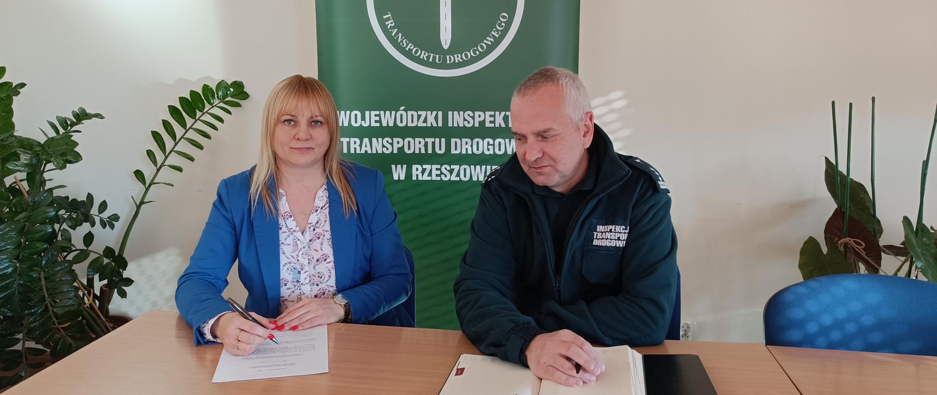 Dwie osoby siedzące przy stole. Kobieta i mężczyzna. Przed nimi leżą dokumenty. Pomieszczenie jest jasne a za nimi stoi zielony baner.