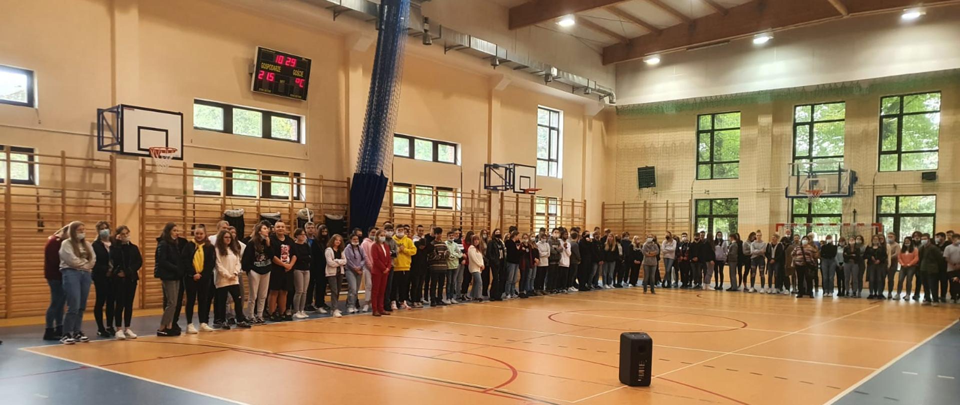 Zdjęcie wykonano w sali gimnastycznej liceum, widok obejmuje obszar parkietu, dwóch ścian i sufitu. Na środku parkietu widać pomarańczowe boisko do siatkówki, a pozostały obszar parkietu jest w kolorze niebieskim. Wzdłóż dwóch boków boiska do siatkówki stoją licealiści, którzy słuchają pogadanki prowadzonej przez strażaka. Na boisku widać również stojący głośnik.