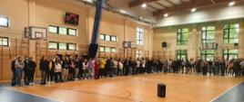 Zdjęcie wykonano w sali gimnastycznej liceum, widok obejmuje obszar parkietu, dwóch ścian i sufitu. Na środku parkietu widać pomarańczowe boisko do siatkówki, a pozostały obszar parkietu jest w kolorze niebieskim. Wzdłóż dwóch boków boiska do siatkówki stoją licealiści, którzy słuchają pogadanki prowadzonej przez strażaka. Na boisku widać również stojący głośnik.