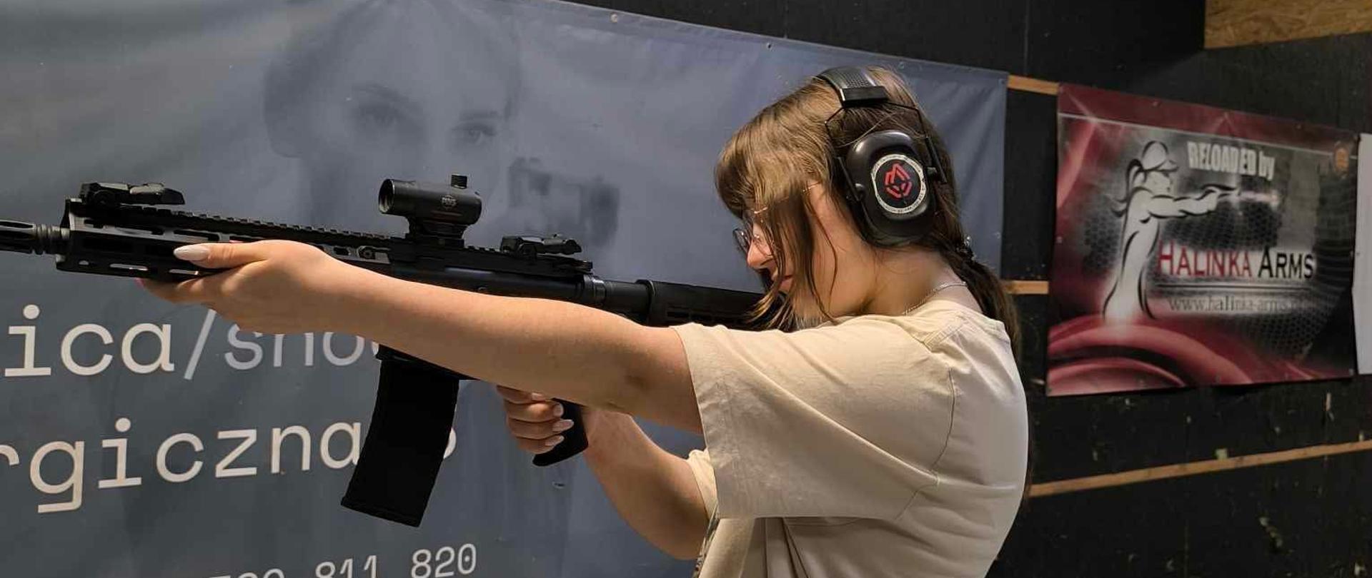 Młoda kobieta stoi na strzelnicy wewnętrznej, celując z karabinka półautomatycznego. Ubrana jest w luźny, beżowy strój i ma na sobie ochronne słuchawki. W tle widoczne są banery reklamowe oraz rura wentylacyjna pod sufitem. Pomieszczenie ma kafelkowaną podłogę i ciemne ściany.