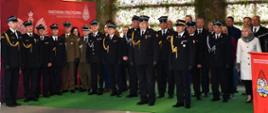 Uroczystość w pomieszczeniu ze ścianą pokrytą wojskową siatką maskującą i podświetleniem. Na pierwszym planie stoi mównica z czerwonym panelem, na którym widnieje logo Państwowej Straży Pożarnej oraz napis "PAŃSTWOWA STRAŻ POŻARNA KOMENDA POWIATOWA PSP W LEŻAJSKU". W tle stoi grupa umundurowanych strażaków w granatowych mundurach wyjściowych PSP oraz kilka osób cywilnych. Na zielonym tle za strażakami widoczny jest duży, czerwony baner z białym logo PSP i napisem "PAŃSTWOWA STRAŻ POŻARNA KOMENDA POWIATOWA PSP W LEŻAJSKU".