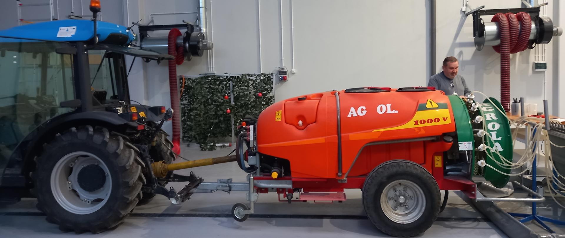 Czerwony opryskiwacz na kołach podłączony do ciągnika w kolorze niebieskim stoi w hali