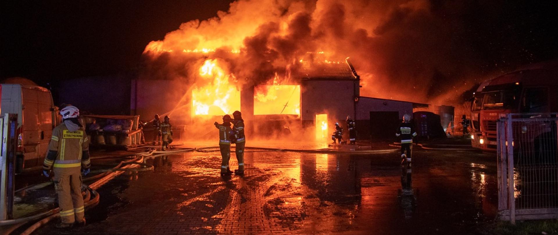 Na zdjęciu budynek objęty w całości pożarem, strażacy ubrani w aparaty powietrzne podają prądy gaśnicze na palący się budynek, po obu stronach palącego się budynku zaparkowane samochody ciężarowe