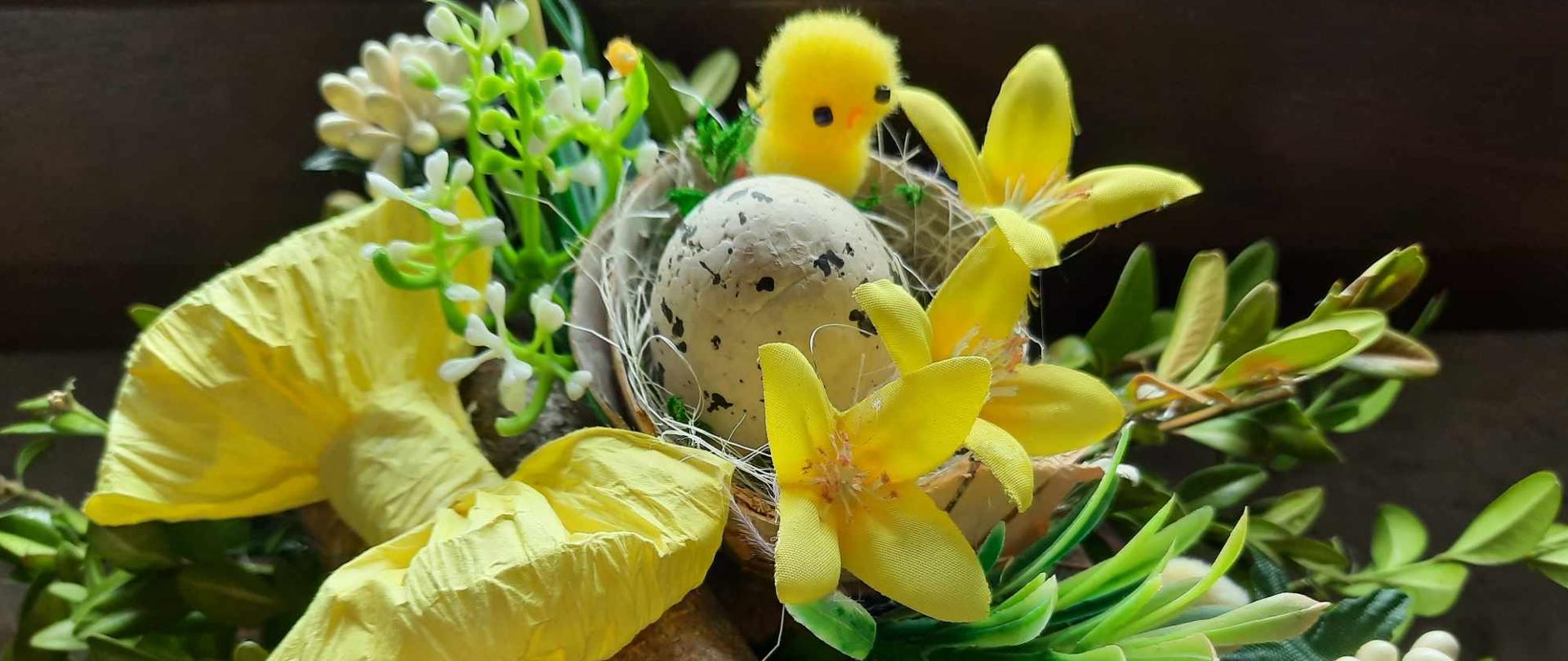 Stroik świąteczny w zielonych, żółtych i kremowych kolorach. Na drewnianym podkładzie umocowane żółta kokarda, gniazdko z pstrokatym jajeczkiem i żółtym kurczakiem, żółtymi kwiatkami, wokół zielone gałązki.