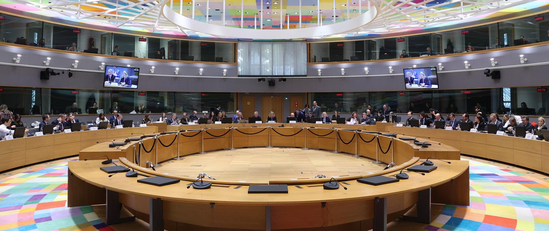 Widok z boku na dużą salę, pośrodku okrągły stół, dookoła niego siedzenia dla wielu osób, nad nim sufit w kolorowe geometryczne wzory.