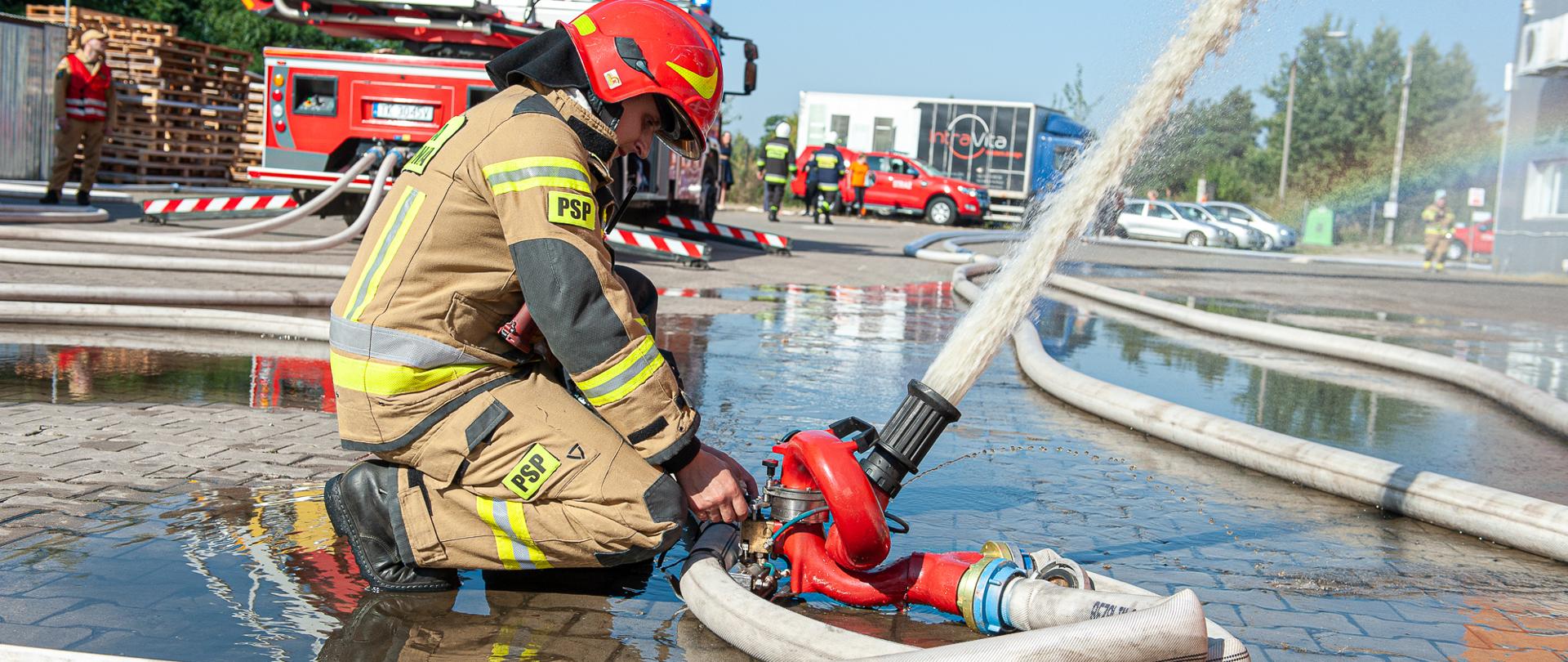 Strażak obsługujący działko wodne na tle samochodu gaśniczego i zakładu.