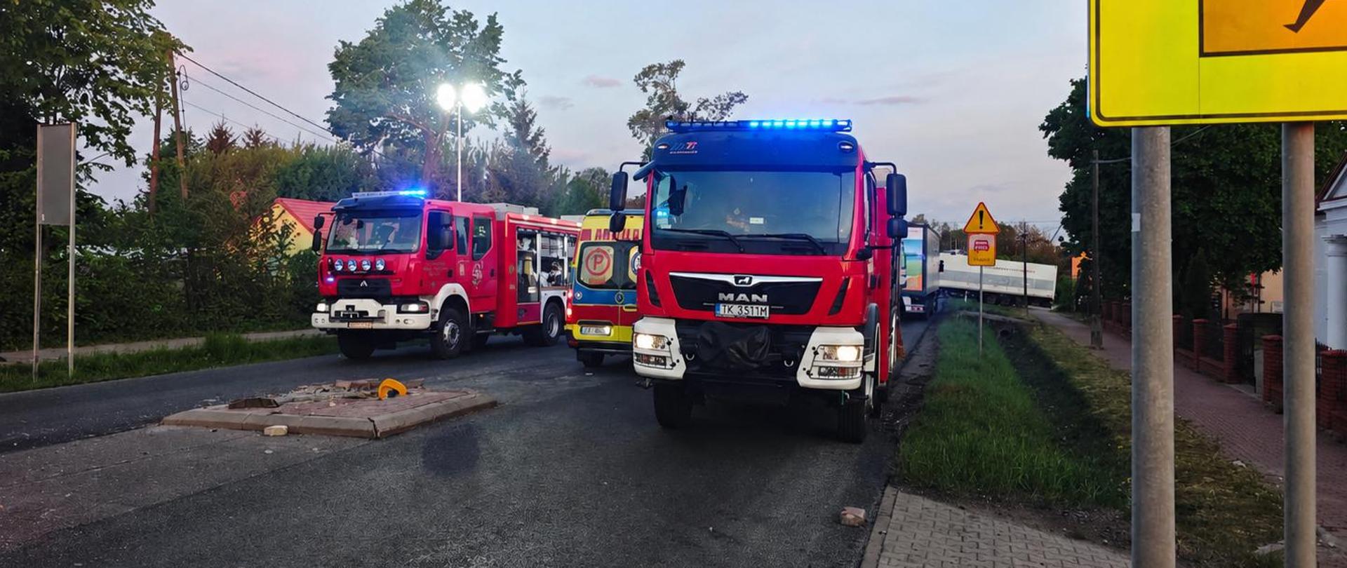Na zdjęciu widać dwa czerwone wozy strażackie stojące na drodze o zmierzchu, z włączonymi niebieskimi światłami alarmowymi. Obok nich znajduje się ambulans oraz ciężarówka. Po prawej stronie widoczny jest chodnik i duży żółty znak drogowy ostrzegający o przejściu dla dzieci. Ulica wygląda na częściowo zablokowaną, a na jezdni znajduje się niewielka wysepka drogowa. W tle widać drzewa, domy i jasne niebo po zachodzie słońca.