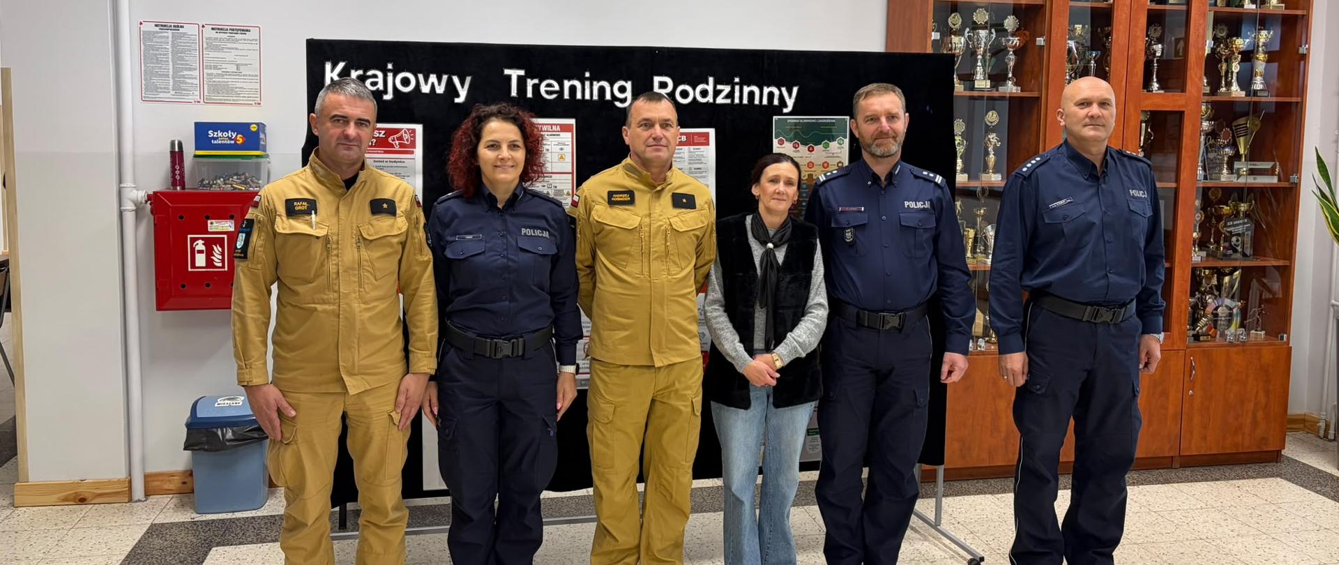Strażacy, policjanci oraz cywilna kobieta pozują wspólnie do pamiątkowego zdjęcia na tle ścianki z napisem „Krajowy Trening Rodzinny”.