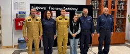 Strażacy, policjanci oraz cywilna kobieta pozują wspólnie do pamiątkowego zdjęcia na tle ścianki z napisem „Krajowy Trening Rodzinny”.