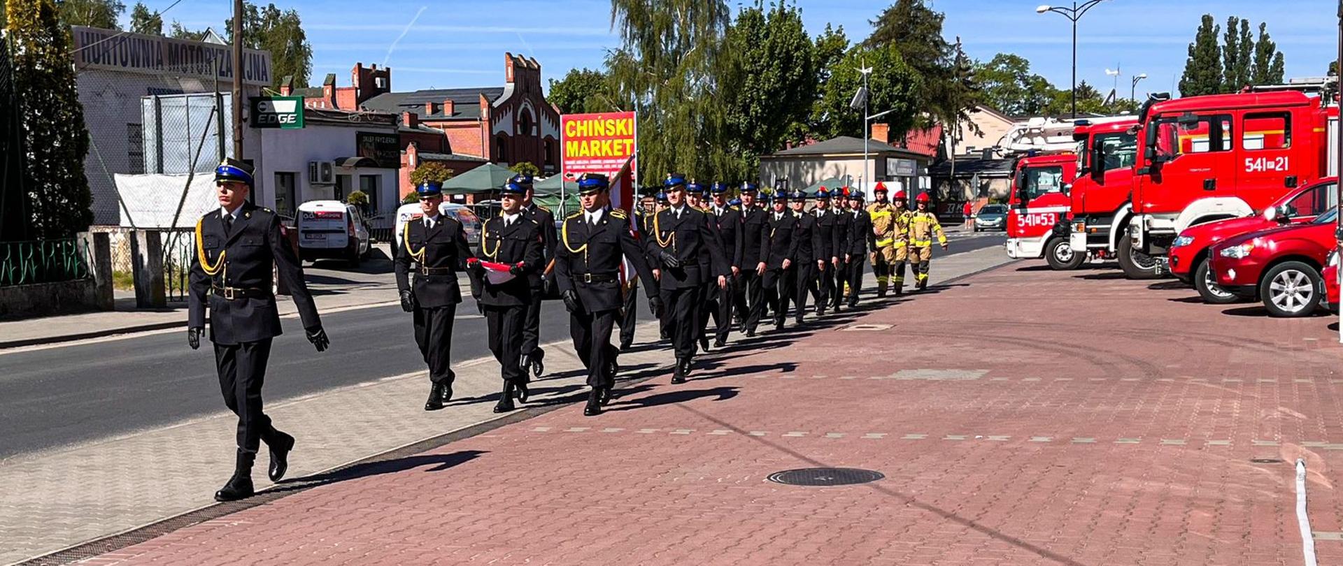 Pododdział strażaków w trakcie wchodzenia na plac, na którym odbywały się obchody dnia strażaka. Po prawej stronie widać samochody strażackie.