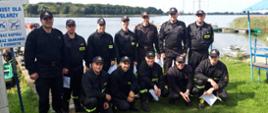 Szkolenia z ratownictwa na obszarach wodnych. Grupa strażaków stoi na trawie i pozuje do wspólnego, pamiątkowego zdjęcia. W tle jezioro i drzewa.