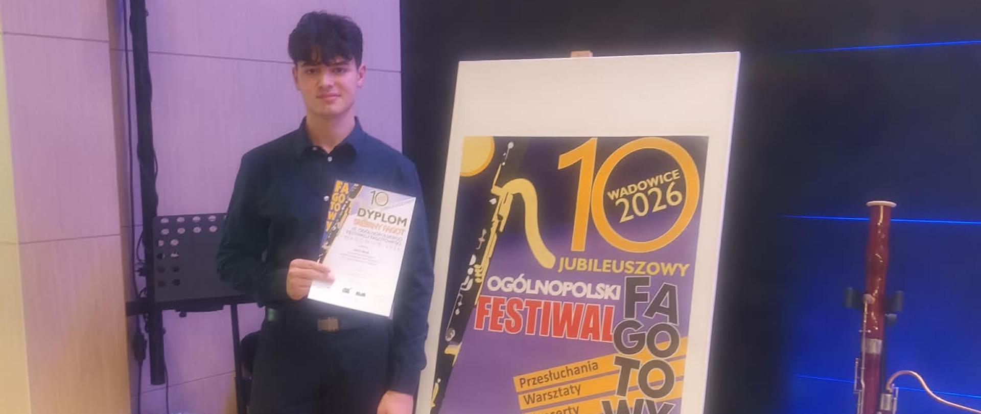 Uczeń stoi na tle plakatu, w ręku trzyma dyplom. Obok plakatu stoi fagot na statywie.