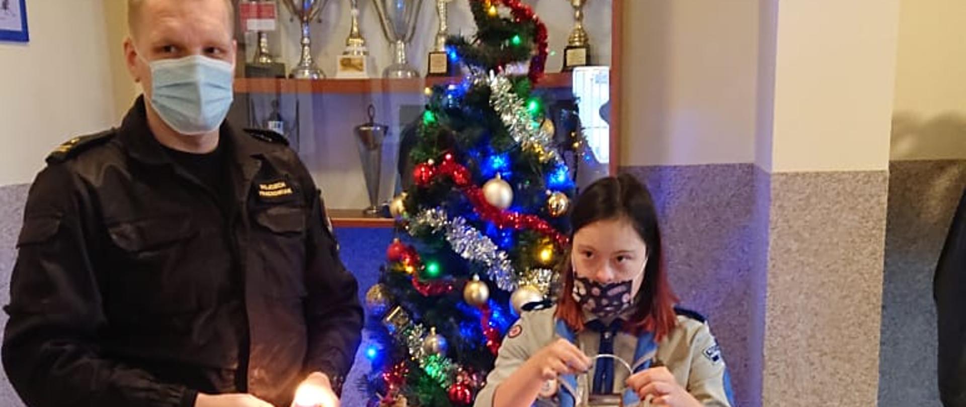 na zdjęciu stoją dwie po lewej mężczyzna w czarnym mundurze PSP trzymający zapalona niewielka czerwoną świeczkę w obu dłoniach, po prawej kobieta ubrana w mundur harcerski trzymająca latarnie ze świeczką. W tle stoi kolorowo przyozdobiona choinka.