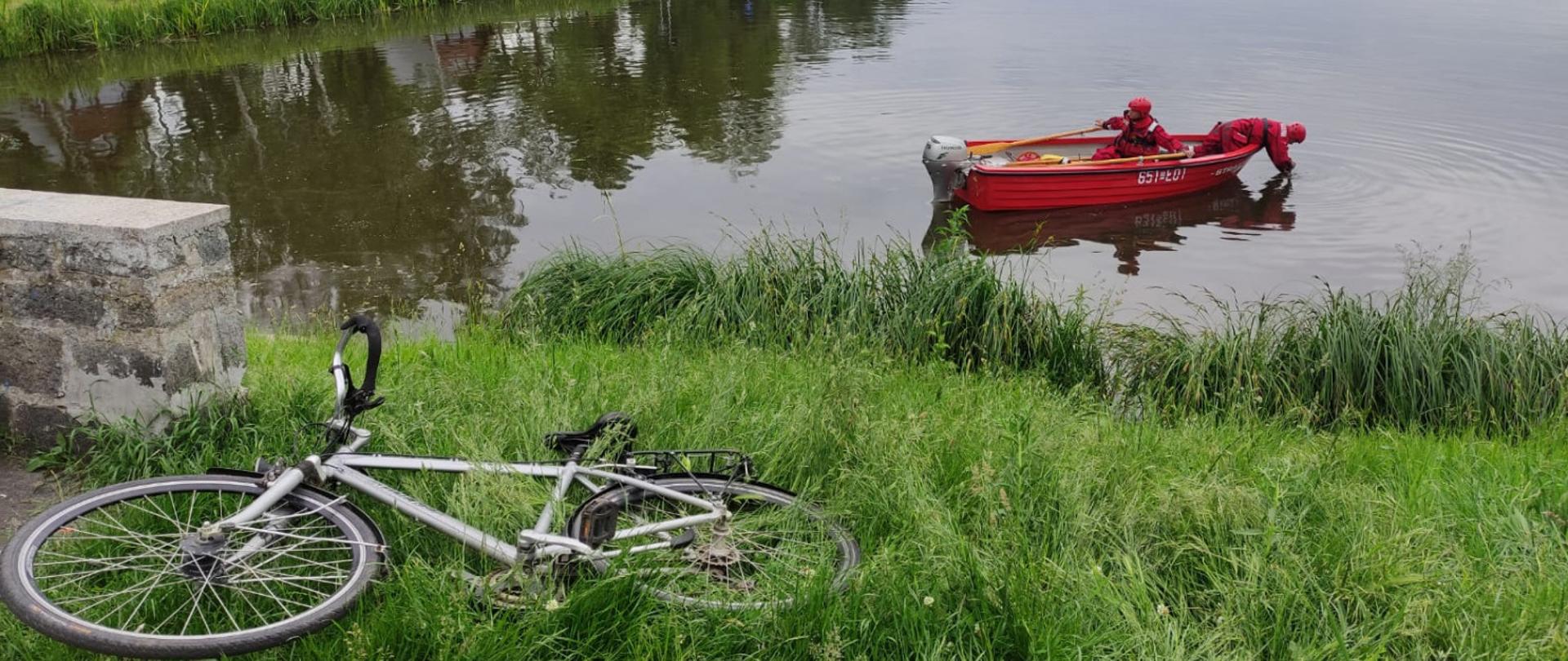 Zdjęcie przedstawia czerwoną łódkę ze strażakami płynąca po jeziorze, na brzegu porzucony rower