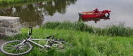 Zdjęcie przedstawia czerwoną łódkę ze strażakami płynąca po jeziorze, na brzegu porzucony rower