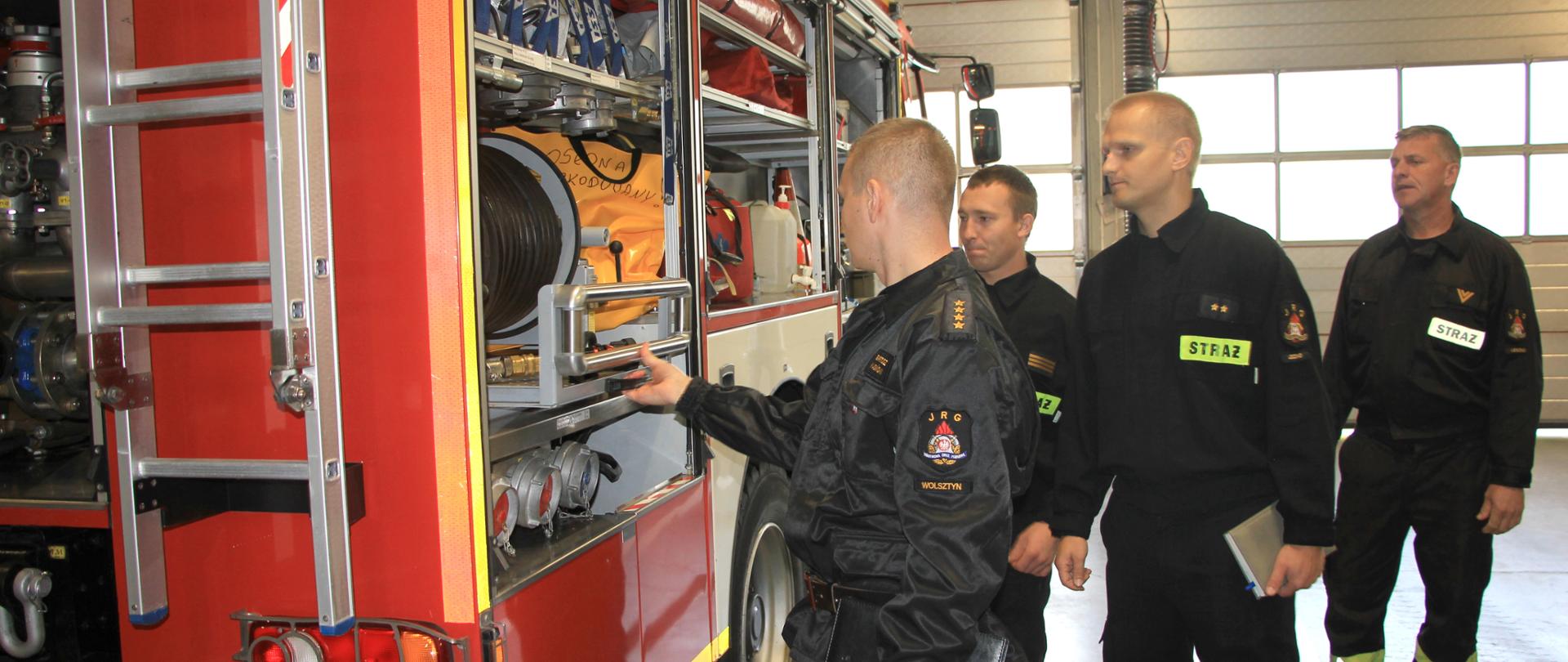 Strażacy w mundurach przeglądają sprzęt i pojazdy pożarnicze stojące w garażu.