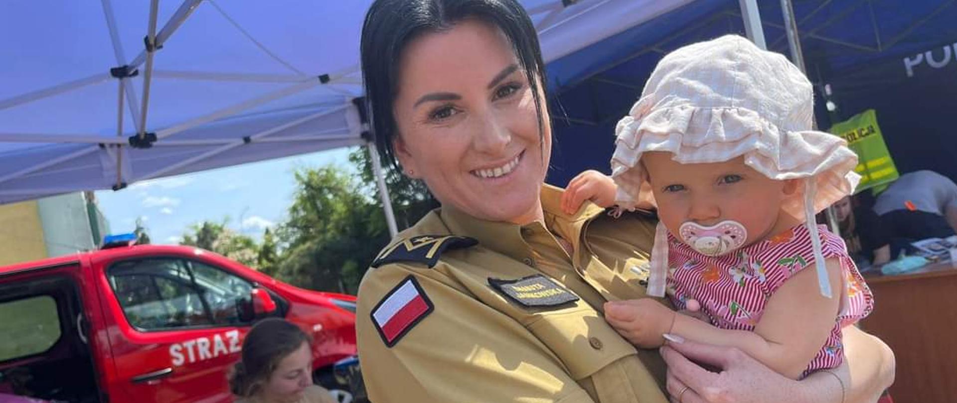 Zdjęcie przedstawia funkcjonariuszkę PSP z dzieckiem na rękach, uśmiechnięte w tle samochód pożarniczy z napisem STRAŻ