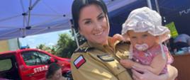 Zdjęcie przedstawia funkcjonariuszkę PSP z dzieckiem na rękach, uśmiechnięte w tle samochód pożarniczy z napisem STRAŻ