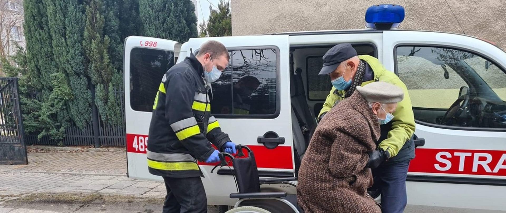 Na zdjęciu widać strażaka oraz mężczyznę pomagających zejść z wózka i wsiąść do samochodu kwatermistrzowskiego ochotniczej straży pożarnej. Kobieta i mężczyzna są spokrewnieni. W tle widać samochód kwatermistrzowski z OSP Skarżysko.
