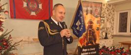 Wnętrze sali wiejskiej. Przed sceną, na sztandaru OSP, flagi Związku OSP RP oraz choinki i prezentów stoi komendant powiatowy PSP w Rawiczu, który trzymając w rękach mikrofon, składa zebranym życzenia świąteczne. Mówca ma na sobie mundur wyjściowy ze sznurem galowym. 