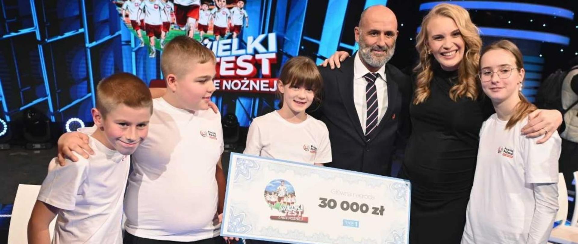 Na zdjęciu trójka dzieci, wśród nich dziewczynka trzymająca prostokątny duży bon na 30000 zł. Obok dzieci dwójka elegancko ubranych dorosłych. W tle grafika przedstawiająca biegnące dzieci i dorosłego - wszyscy ubrani w narodowe stroje piłkarskie.