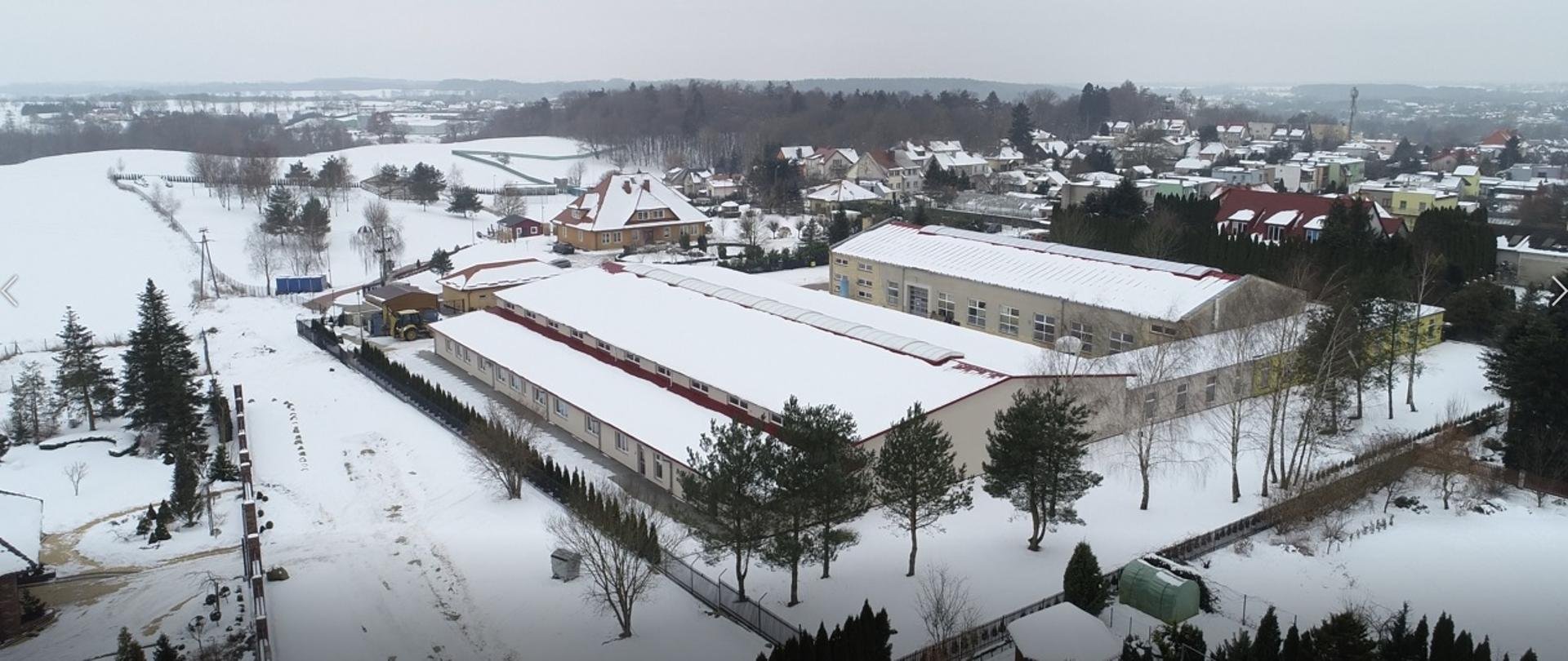 Zdjęcie przedstawia budynek wielkopowierzchniowy z lotu ptaka. dach budynku jest pokryty śniegiem. W oddali widzimy budynki mieszkalne, lasy i pola.