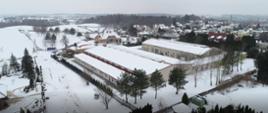 Zdjęcie przedstawia budynek wielkopowierzchniowy z lotu ptaka. dach budynku jest pokryty śniegiem. W oddali widzimy budynki mieszkalne, lasy i pola.