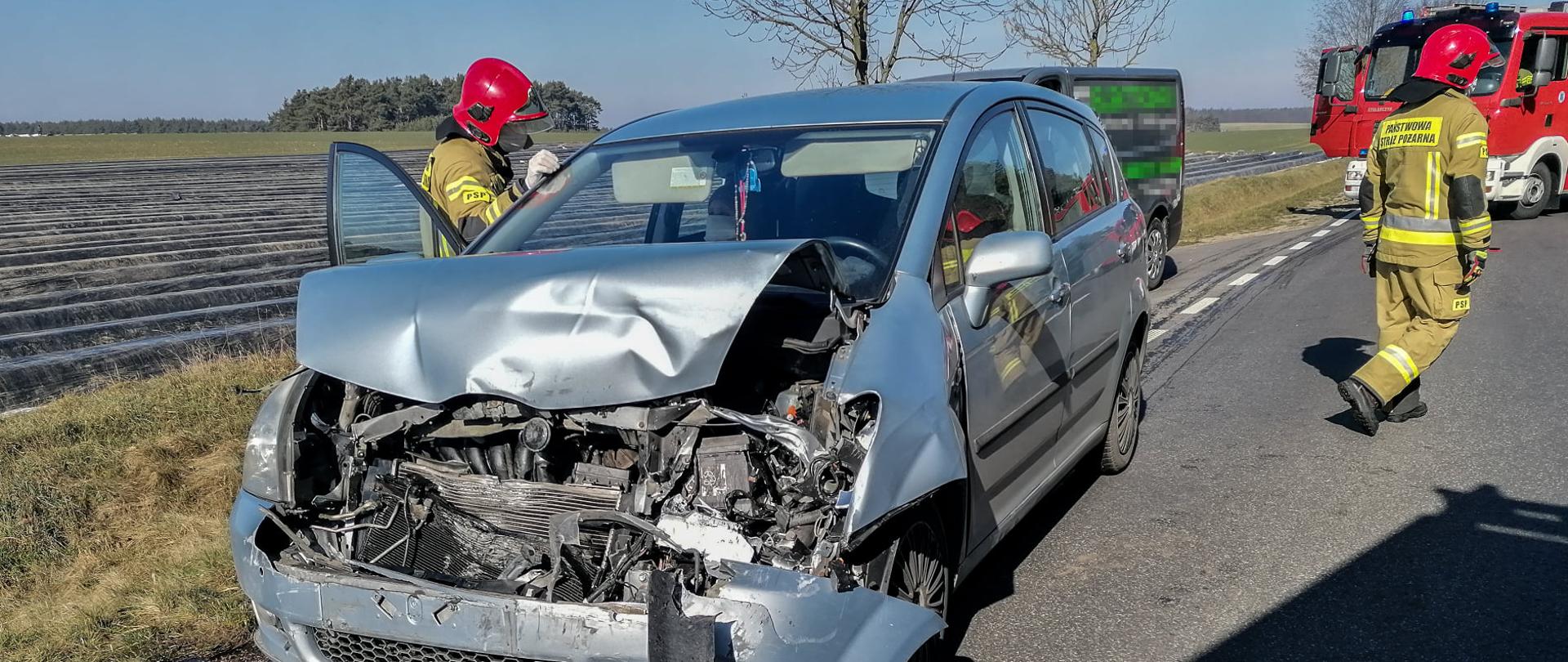Rozbity samochód na drodze. Dwóch strażaków odwróconych tyłem, w tle samochód pożarniczy.