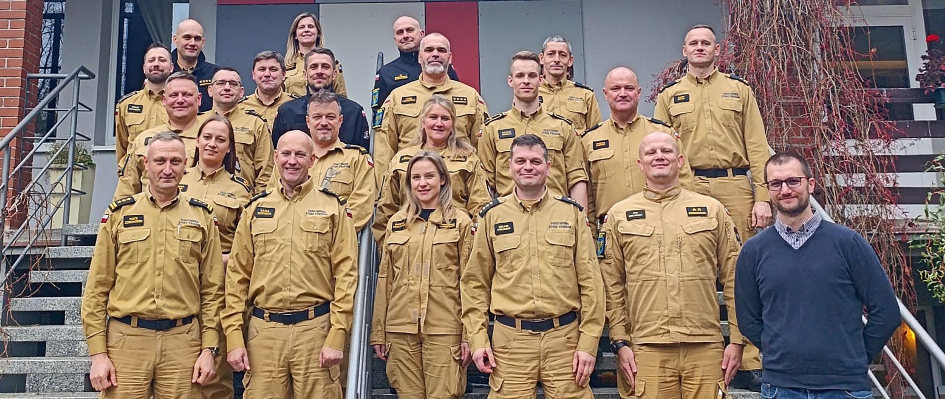 Dwudziestoosobowa grupa funkcjonariuszy i pracowników PSP województwa opolskiego pozuje do zdjęcia grupowego na schodach przed budynkiem w Turawie. Dziewiętnaście osób w mundurach PSP, jedna w stroju cywilnym. Zdjęcie wykonano podczas narady szkoleniowej w dniach 20–21 listopada 2025 roku.