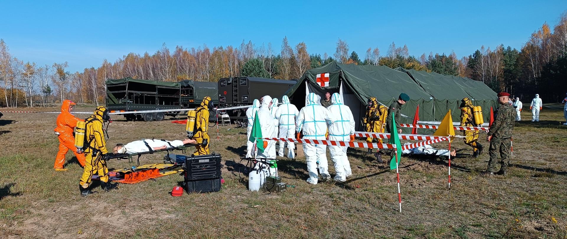 Ratownicy w kombinezonach na tle namiotu i samochodów wojskowych