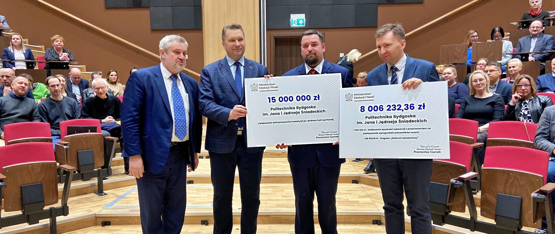 Minister stoi obok trzech mężczyzn w garniturach, dwaj z nich trzymają wielkie symboliczne czeki z napisami 15 000 000 zł i 8 006 232,36 zł.