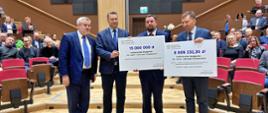 Minister stoi obok trzech mężczyzn w garniturach, dwaj z nich trzymają wielkie symboliczne czeki z napisami 15 000 000 zł i 8 006 232,36 zł.