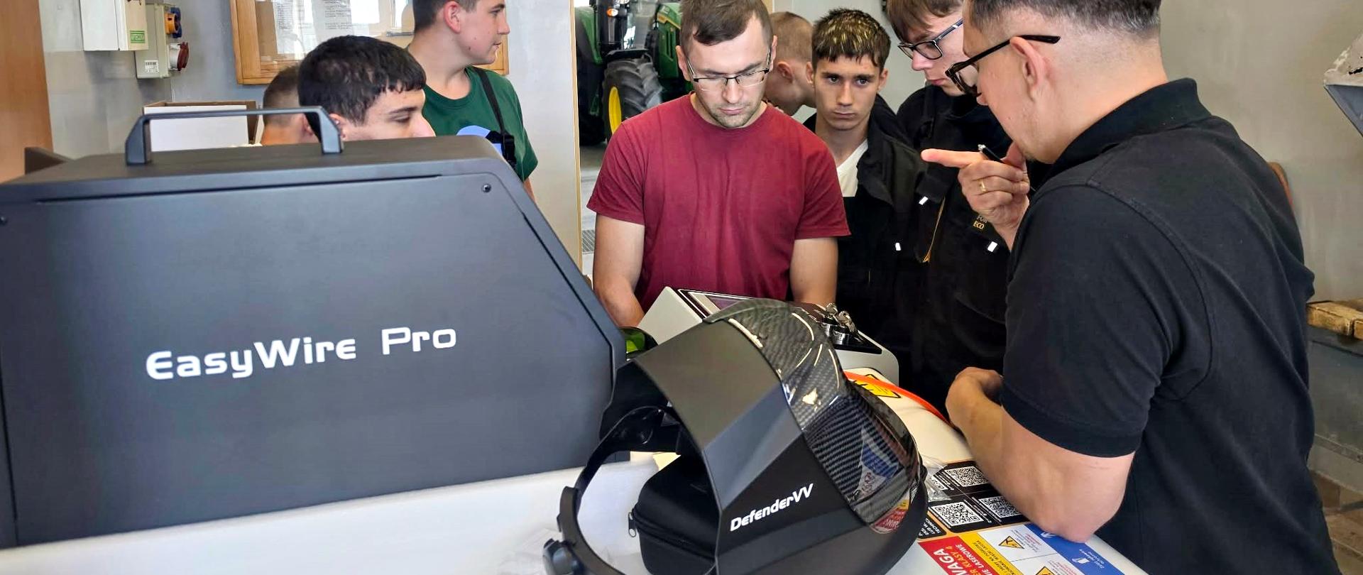 Zdjęcie przedstawia szkolenie dotyczące technologii spawania laserowego. Kilka osób zgromadzonych wokół stanowiska roboczego obserwuje prezentację urządzenia oznaczonego napisem „EasyWire Pro”. Na stole, obok sprzętu, znajduje się przyłbica spawalnicza z oznaczeniem „DefenderVV”.
