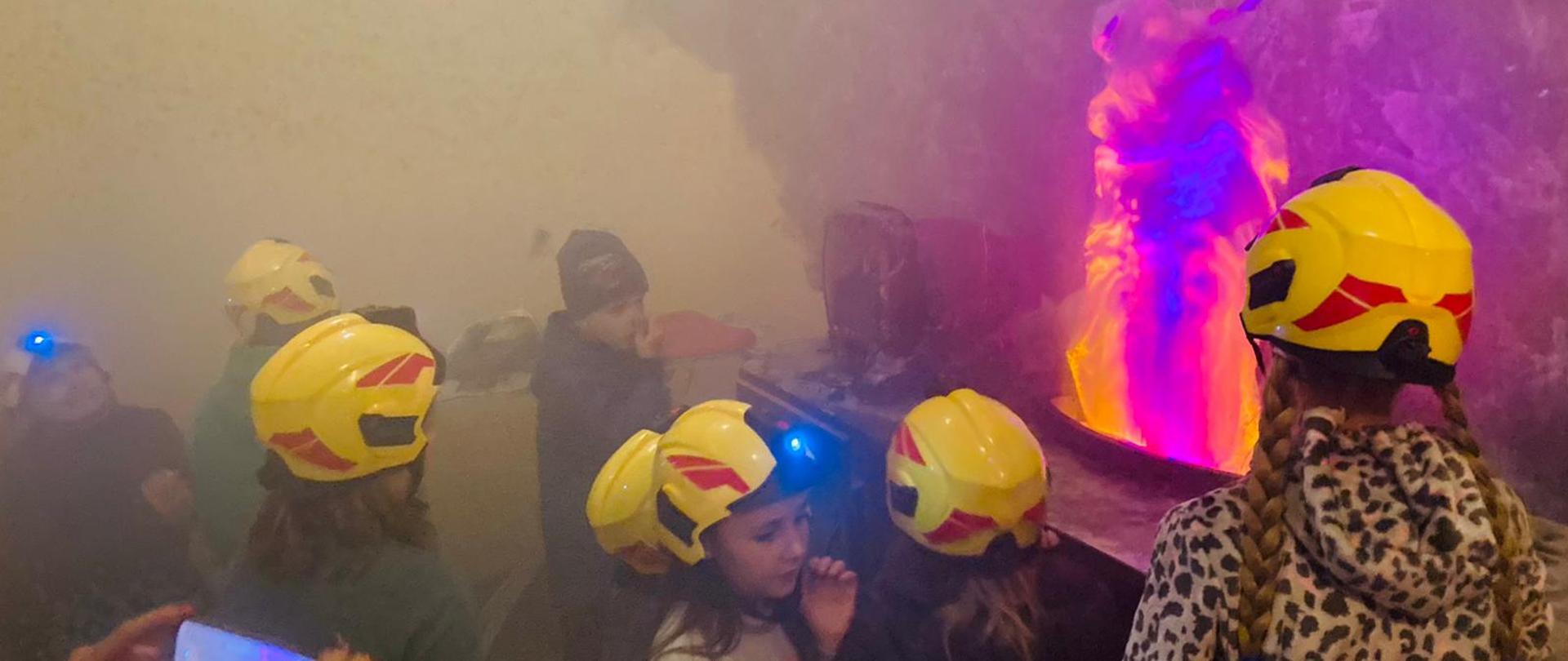 Zdjęcie przedstawia dzieci ubrane w zabawkowe żółte kaski, w pomieszczeniu kuchni symulującej pożar i zadymienie