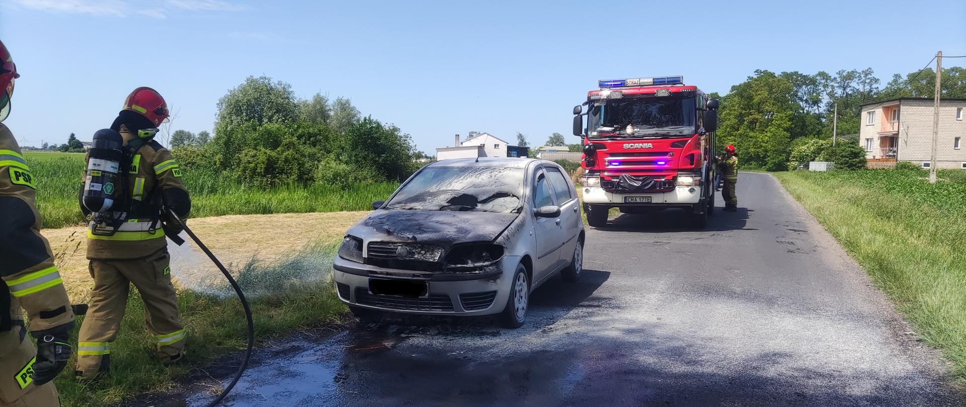 Zdjęcie na pierwszym planie przedstawia strażaków Państwowej Straży Pożarnej ubranych w ubrania koloru piaskowego oraz aparaty ochrony układu oddechowego, podczas prowadzenia działań ratowniczo-gaśniczych pożaru samochodu. na drugim planie samochód osobowy ze spaloną komorą silnika oraz przednią szybą. Na kolejnym planie wóz straży pożarnej koloru czerwonego.