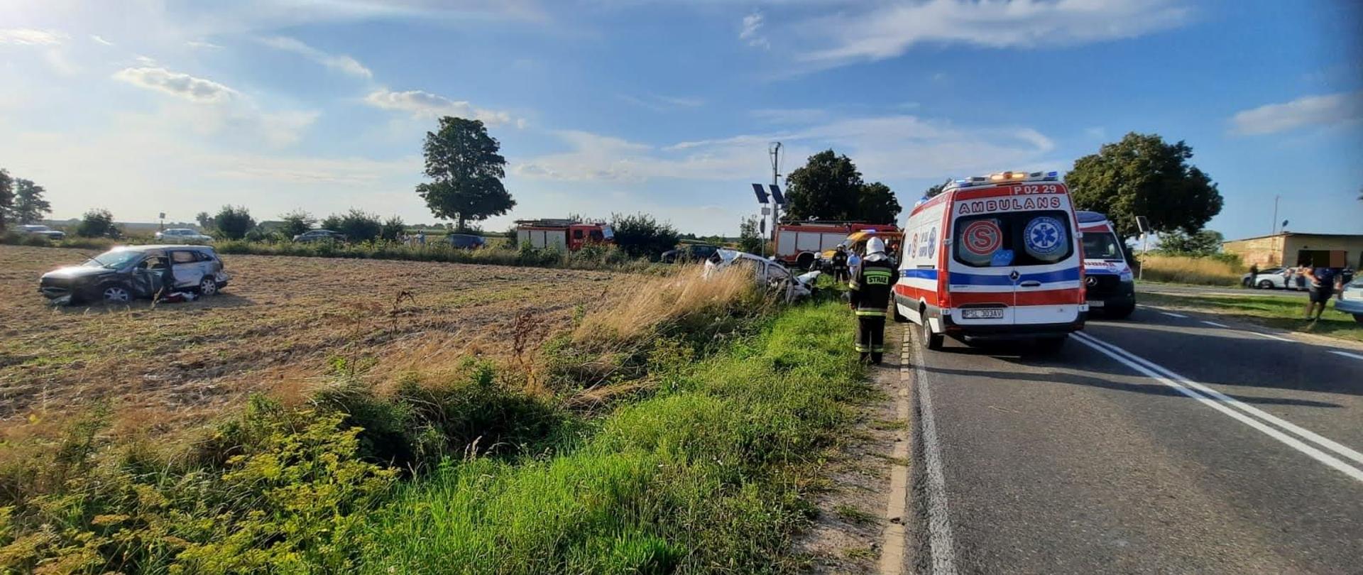 Na pierwszym planie karetki pogotowia ratunkowego obok stoi tyłem strażak po lewej stronie na polu uprawnym rozbity srebrny samochód oraz biały znajdujący się w rowie w oddali czerwone samochody pożarnicze oraz inne samochody stojące na drodze. Po prawej stronie przy żółtym budynku stoją jakieś osoby obok widać samochody na horyzoncie drzewa 
