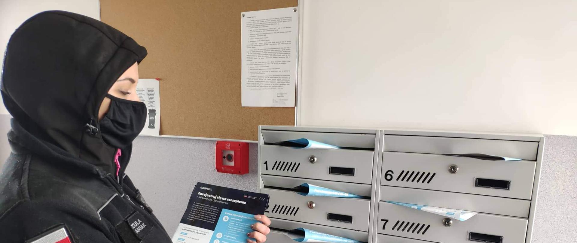 Umundurowana druhna OSP Ośno Lubuskie stojąca z ulotkami przy skrzynkach pocztowych na klatce schodowej. W skrzynkach już są widoczne umieszczone ulotki.