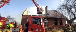 Na pierwszym planie zdjęcia widać podnośnik hydrauliczny straży pożarnej z rozłożonym koszem w kierunku budynku objętego pożarem. Połowa dachu budynku uległa spaleniu i zawaleniu. Na zdjęciu widać pracujących strażaków.