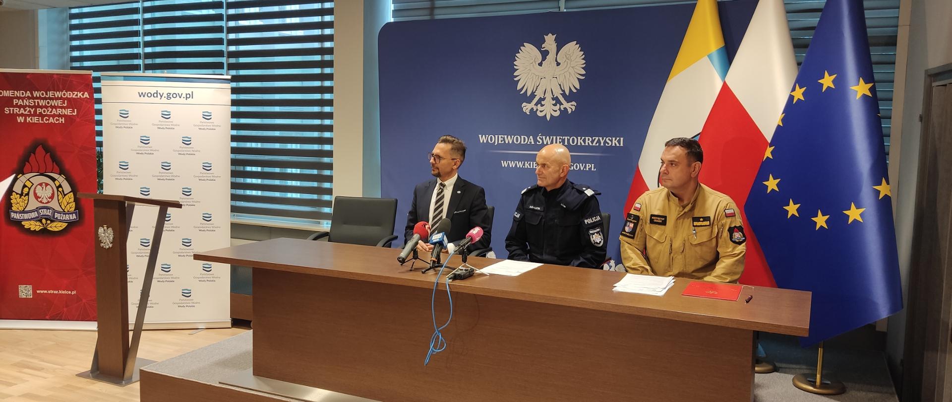 Na zdjęciu widzimy 3 mężczyzn siedzących za stołem prezydialnym. siedzą od lewej Mężczyzna w garniturze, a od prawej dwóch mężczyzn w mundurach (pierwszy od prawej w piaskowym mundurze Państwowej Straży Pożarnej a drugi to granatowy mundur Policji)