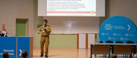 Komendant Centralnej Szkoły PSP przemawia do mikrofonu trzymanego w dłoni stojąc na podwyższeniu w czasie konferencji. W głębi ekran multimedialny z wyświetlonym slajdem prezentacji
